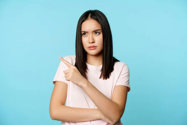 Angry asian woman sulking, frowning upset, pointing fingers at upper left corner, standing in t-shirt over blue background — Stock Photo, Image