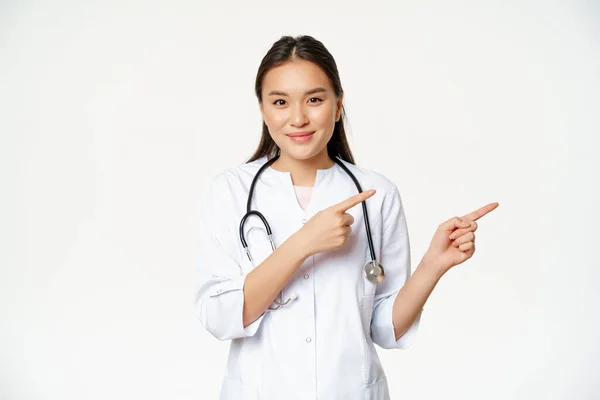 Zdravotnictví, medicína. Usmívající se asijská lékařka, ukazující prstem vpravo, zobrazující inzerát, nemocniční informace, stojící nad bílým pozadím — Stock fotografie