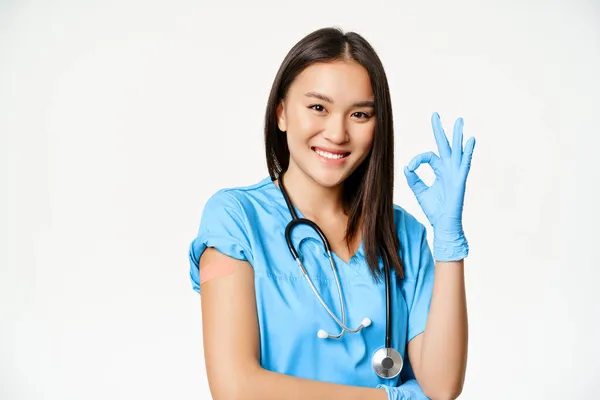 Leende sjuksköterska, asiatisk kvinnlig läkare i buskar, visar okej tecken och vaccinerad arm med medicinskt gips, rekommenderar vaccination från covid-19, vit bakgrund — Stockfoto