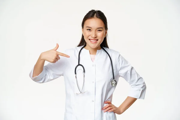 Sorrindo asiático médico feminino, real profissional médico trabalhador, apontando os dedos para si mesma, vestindo roupão médico e estetoscópio, fundo branco — Fotografia de Stock