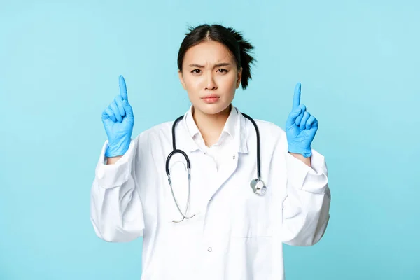 Naštvaná a znepokojená doktorka, lékař ukazuje prsty nahoru, zamračeně se mračí, stojí v lékařské uniformě nad modrým pozadím — Stock fotografie