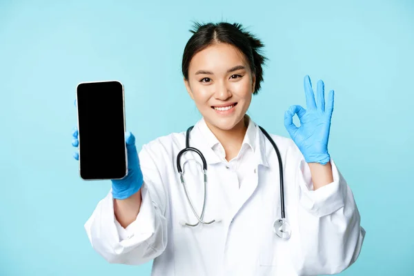 Web healthcare, concepto de ayuda en línea. Confiado médico o enfermera asiática, mostrando la pantalla del teléfono inteligente, interfaz de la aplicación y signo bien en la aprobación, recomendando sitio web — Foto de Stock