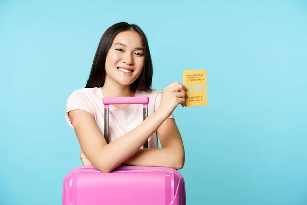 Happy usměvavá asijská dívka, turistické stánky s roztomilým kufrem na letišti, ukazuje mezinárodní covid-19 očkovací průkaz pro cestující, bezpečné cestování po očkování, modré pozadí — Stock fotografie