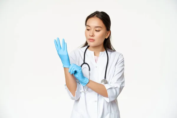 Portré nő ázsiai orvos felvesz gumikesztyűt, hogy vizsgálja meg a beteg a klinikán, álló egészségügyi egyenruha fehér háttér — Stock Fotó