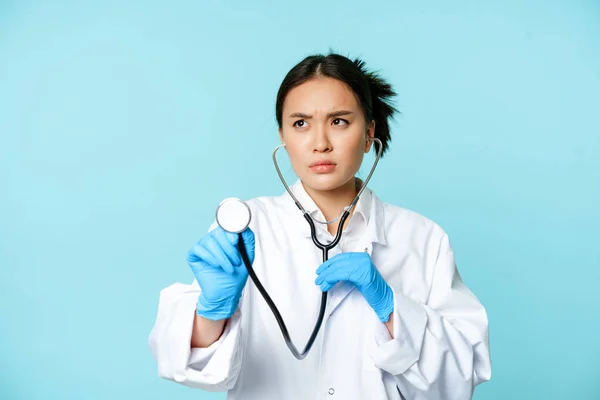 Ciddi bir tıp çalışanı görüntüsü, Asyalı doktor steteskopla hastanın akciğerlerini dinliyor, kişiyi inceliyor, mavi arka planda duruyor. — Stok fotoğraf