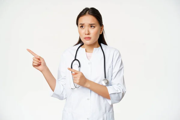 Şüpheci kadın doktor, endişeli hemşire endişeli bir yüz ifadesiyle sol tarafa bakıyor, beyaz tıbbi bornozla stüdyonun arka planında duruyor. — Stok fotoğraf