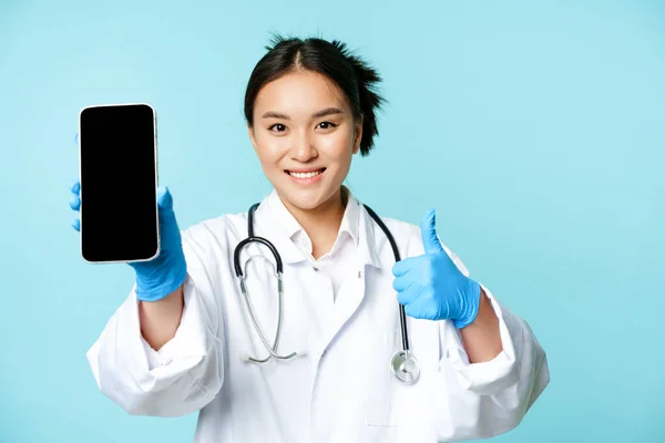Web healthcare, concepto de ayuda en línea. Sonriendo asiática médico o enfermera, mostrando la pantalla del teléfono inteligente, interfaz de la aplicación y pulgares hacia arriba, recomendando sitio web — Foto de Stock