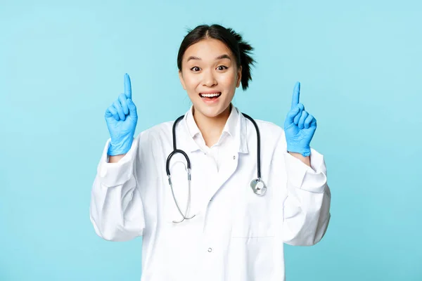 Hevesli Asyalı doktor, kadın doktor parmaklarını kaldırıp gülümsüyor, reklam gösteriyor, tanıtım yapıyor, mavi arka plan. — Stok fotoğraf