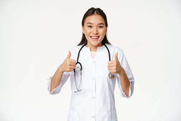 Usmívající se asijské žena lékař, na sobě lékařské roucho, ukazující palce nahoru potěšen, doporučující zdravotní produkt, stojící nad bílým pozadím — Stock fotografie