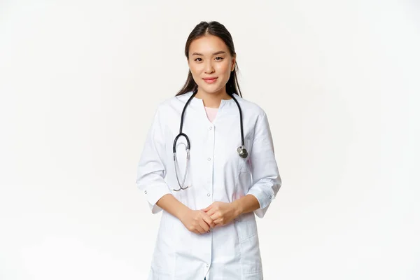 Usmívající se asijský zdravotnický pracovník se stetoskopem, nosí lékařskou uniformu, vypadá užitečné na pacienta, stojící nad bílým pozadím — Stock fotografie