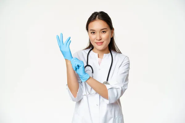 Usmívající se sebevědomý asijský lékař dal na lékařské rukavice, připravuje se na vyšetření pacienta, stojí v uniformě nad bílým pozadím — Stock fotografie
