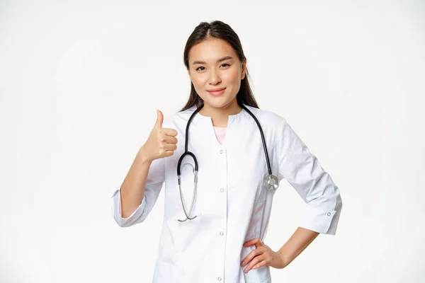 Usmívající se lékař v lékařské uniformě ukazuje palce nahoru. Příjemná asijská žena lékař potvrzuje smth, doporučující produkt, bílé pozadí — Stock fotografie