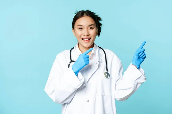 Boldog mosolygós nővér, ázsiai női orvos mutatva jobbra, bemutató promo, klinika hirdetés, álló orvosi egyenruha kék háttér — Stock Fotó