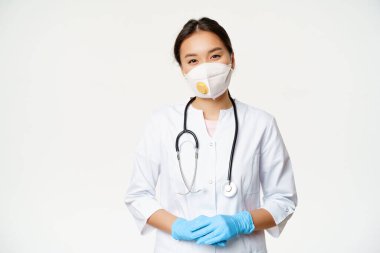 Solunum cihazında gülümseyen Asyalı kadın doktor, tıbbi maske ve lastik eldivenler, yardıma hazır, beyaz arka planda klinik üniforması içinde duruyor. Covid-19 ve karantina konsepti