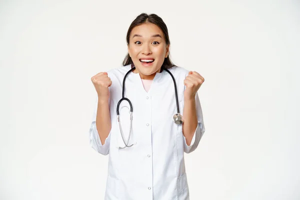 Hevesli Asyalı kadın doktor kutluyor, kamerada umut dolu ve mutlu görünüyor, zafer kazanıyor, beyaz arka plana karşı tıp üniforması içinde duruyor. — Stok fotoğraf