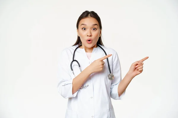 Surprised female doctor say wow, pointing right and looking amazed with discounts in clinic, showing way, standing in medical uniform against white background — Stock Photo, Image
