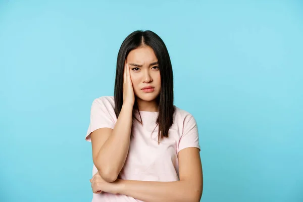 Беспокойная молодая азиатская женщина, держа руку за голову, хмурясь и глядя с недовольным, обеспокоенным лицом, стоящая в футболке на синем фоне — стоковое фото
