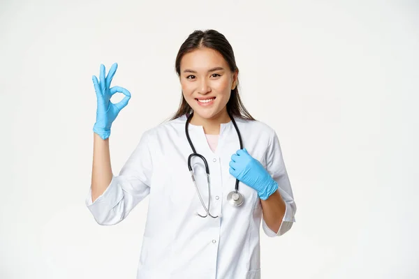 Usmívající se zdravotnice, asijská žena lékař v gumových rukavicích a lékařské uniformě, ukazuje souhlas, dobře znamení, bílé pozadí — Stock fotografie