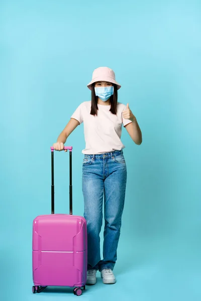 Vertikální záběr očkované asijské dívky v lékařské masce obličeje, na dovolené, stojící s kufrem a ukazující palec nahoru, modré pozadí — Stock fotografie