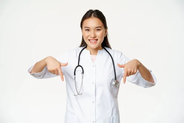 Sorrindo asiático mulher médico, apontando os dedos para baixo e mostrando negócio promo, descontos no serviço de clínica, anúncio hospitalar, fundo branco — Fotografia de Stock
