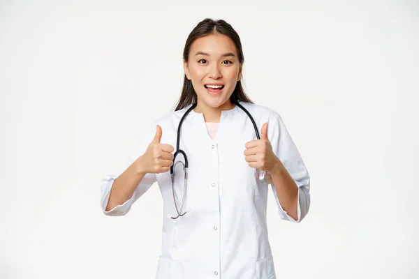 Feliz sorrindo asiático mulher médico, mostrando polegares para cima na aprovação, satisfeito com a condição do paciente, aprovar e similares, de pé sobre fundo branco — Fotografia de Stock