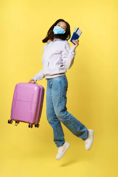 Full length shot šťastný asijské žena jumping s kufrem a vstupenky na turné, držení pasu, jede na dovolenou, stojící nad žlutým pozadím — Stock fotografie