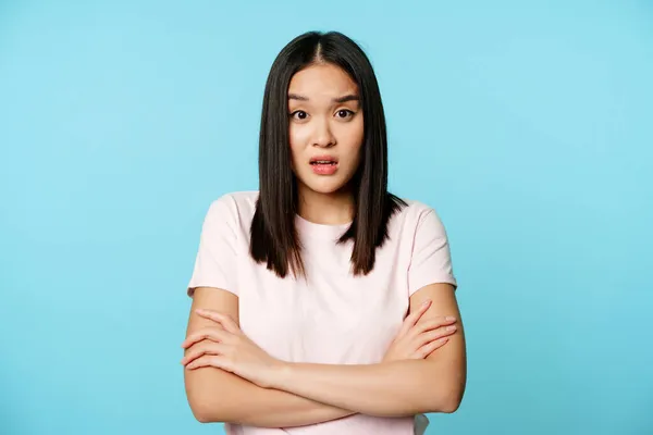 Портрет молодої азіатки, що слухає з заплутаним, шокованим виразом обличчя, стоїть у футболці на синьому фоні — стокове фото