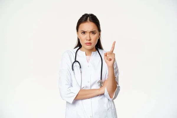 Doktor projevuje nesouhlas, nadává pacientovi. Rozzlobený asijský lékař potřásá prstem a svraštělým obočím, zklamán špatným chováním, stojí nad bílým pozadím — Stock fotografie