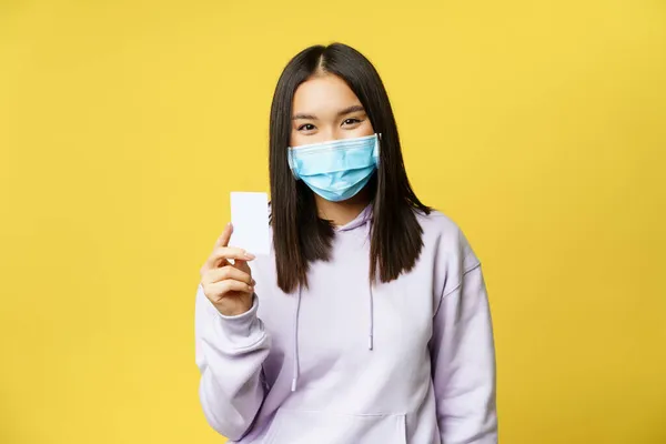 Asiatisk kvinna i medicinsk ansiktsmask, visar kreditkort, passera, står över gul bakgrund — Stockfoto