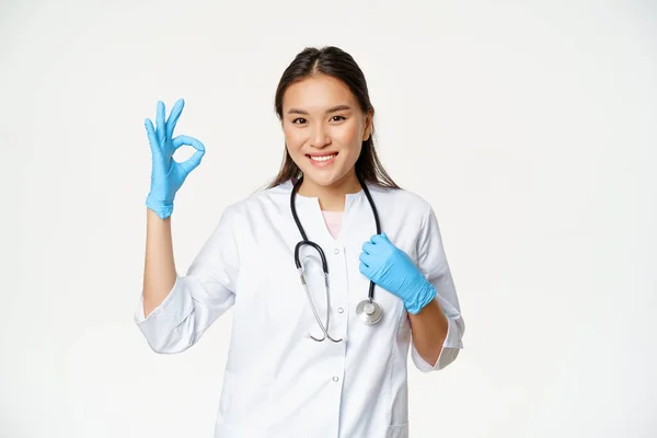 Sonriente trabajador de la salud, mujer asiática médico en guantes de goma y uniforme médico, muestra aprobación, signo de bien, fondo blanco Imagen De Stock