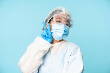 Mutlu, kawaii Asyalı kadın doktor, kişisel koruyucu ekipman hemşiresi, V-işareti barış jesti gösteren yüz maskesi, mavi arka plan