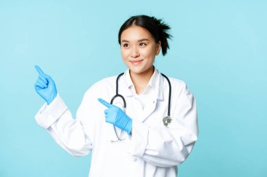Gülümseyen Asyalı doktor, üniformalı sağlık görevlisi, fotokopi alanını işaret edip sola bakıyor, mavi arka planda duruyor.