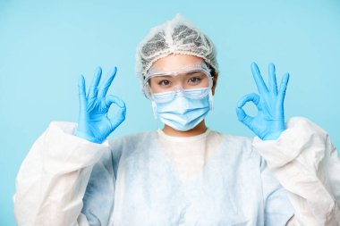 - Tamam. Gülümseyen Asyalı kadın doktor, kişisel koruyucu ekipman hekimi, tamam işareti gösteriyor, mavi arka planda duruyor.