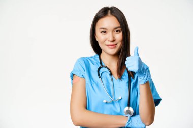 Gülümseyen hemşire, önlüklü Asyalı kadın doktor, baş parmaklarını gösteriyor ve koluna tıbbi alçı takılmış, covid 19 aşısı öneriyor, beyaz arka plan.