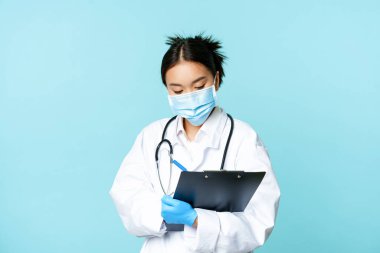 Koreli doktor, tıbbi maske takan kadın hemşire hasta bilgilerini yazıyor, hastane vardiyasını inceliyor, mavi arka planda üniforma içinde duruyor.