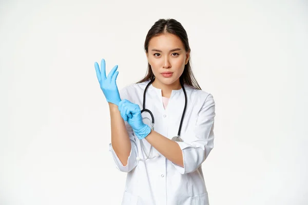 Sebevědomá ošetřovatelka, lékař nasadit gumové lékařské rukavice pro klinické vyšetření pacienta, stojící vážně v uniformě zdravotnického pracovníka, bílé pozadí — Stock fotografie