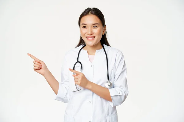 Sorrindo feminino asiático médico em uniforme médico, apontando os dedos e olhando para a esquerda na propaganda, espaço promo cópia, de pé em roupão contra fundo branco — Fotografia de Stock