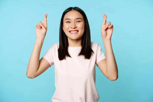 Entusiástico asiático menina desejando, cruz dedos para boa sorte e sorrindo, espreitando para câmera esperançosa, de pé sobre fundo azul — Fotografia de Stock