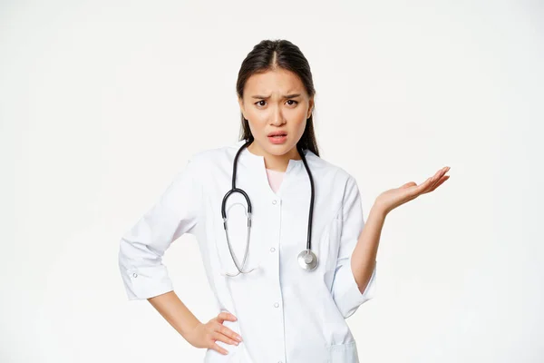混乱したアジアの女性医師は、患者を理解することはできません,困惑し、いらいら見て,疑問のポーズを手を上げます,白い背景の上に医療制服に立って — ストック写真