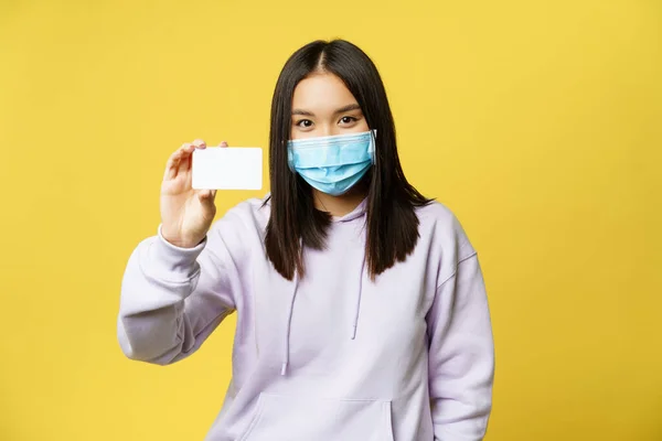 Smiling korean girl in face mask, showing card, standing in casual hoodie over yellow background — Stock Photo, Image