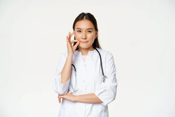 Physician-patient privilege, medical confidentiality. Asian woman doctor showing silence, mouth zip gesture, secret, standing over white background — Stock Photo, Image