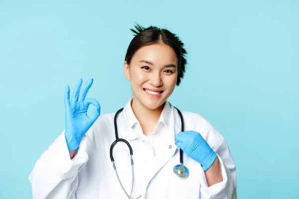 Usmívající se asijský lékař, ženský lékař dotýkající se stetoskopu, ukazuje v pořádku, ok znamení v souhlasu, modré pozadí — Stock fotografie