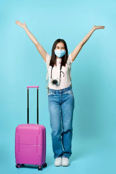 Dlouhý záběr šťastné asijské dívky turista, na obličeji lékařské masky, stojící v blízkosti roztomilý kufr, jede na dovolenou během covid pandemie, nadšený z cestování do zahraničí — Stock fotografie