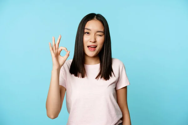 Satisfecho chica asiática guiña y muestra ok signo, dar aprobación, recomienda, se para en camiseta sobre fondo azul —  Fotos de Stock
