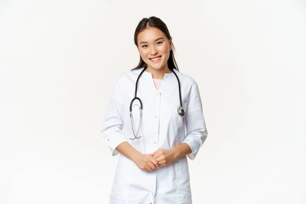 Usmívající se asijská žena lékař, na sobě lékařské roucho a stetoskop, vypadá příjemně na pacienta, stojí nad bílým pozadím — Stock fotografie