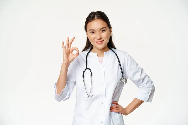 Velmi dobře. Usmívající se sebevědomý asijský lékař, ukazuje v pořádku, ok znamení v souhlasu, potvrdit smth, říká ano, dát souhlas, stojící v lékařské uniformě, bílé pozadí — Stock fotografie