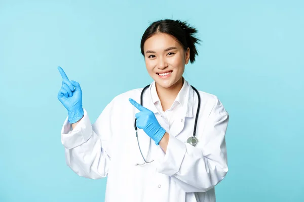 Sorrindo asiático médico feminino, terapeuta apontando os dedos no canto superior esquerdo, mostrando propaganda médica, de pé sobre fundo azul — Fotografia de Stock