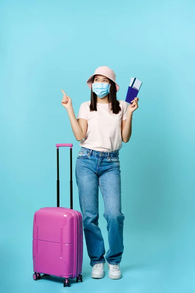 Usmívající se asijský cestovatel, turista dívka v masce obličeje, stojící v blízkosti kufru se dvěma jízdenkami a cestovním pasem, ukazující stranou přemýšlivý, výběr prázdninového cíle, modré pozadí — Stock fotografie