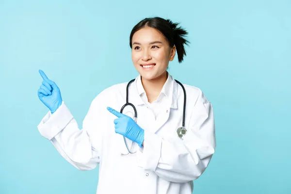 Asiática sonriente médico, médico trabajador de la salud, señalando los dedos a la izquierda, buscando complacido en la publicidad, producto publicitario, fondo azul — Foto de Stock