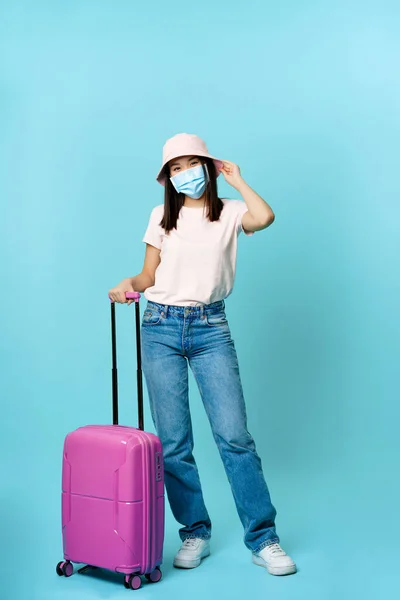 Plná délka portrét šťastné asijské ženy, cestovatel s kufříkem s maskou lékařské tváře během covid pandemie, jede na dovolenou, modré pozadí — Stock fotografie